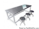The image shows a long, rectangular table with four stools attached to it. The table is made of stainless steel and has a shi...