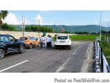 The image shows a group of people standing on the side of a road, waiting to board a car. There are three cars parked on the...