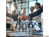 The image shows two men shaking hands in an office setting. The man on the left is wearing a suit and tie and is holding the...