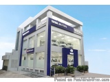 The image shows the exterior of a Tata Motors dealership. The dealership is a modern building with a white exterior and blue...