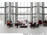 The image shows a large open space with large windows that offer a view of the city. The space is filled with people sitting...