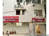 The image shows the entrance of a building named Sai Shreyas Residency. The building is white with a red signboard that reads...